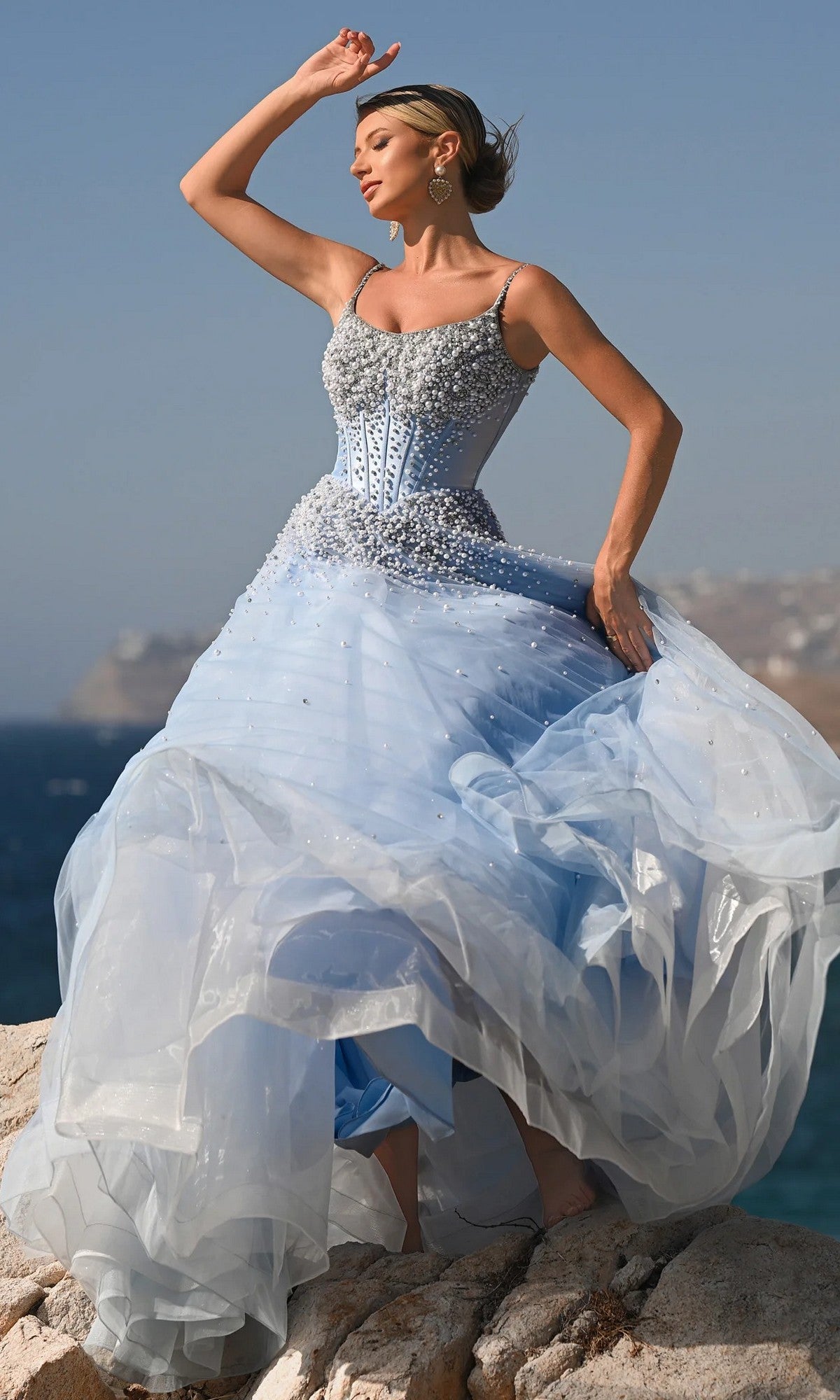 A woman poses on a rocky seaside cliff in the Portia and Scarlett PS26208 corseted ball gown, light blue with embellishments, its tulle skirt and her hair flowing in the wind under a clear sky.